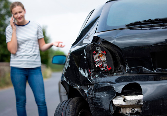 Woman on the phone beside a car with severe rear-end damage and broken taillight after a collision, illustrating insurance evaluation and Diminished Value Claim California following an auto accident
