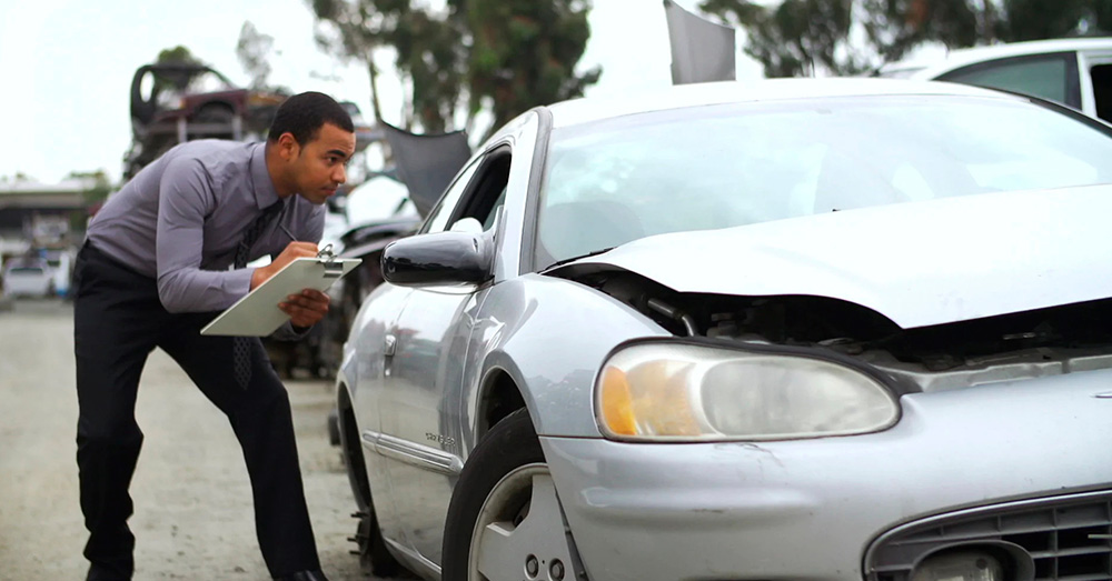 Auto insurance adjuster inspecting front-end damage on a silver sedan at a salvage yard, documenting loss for valuation and Diminished Value Claim Maine after an accident.