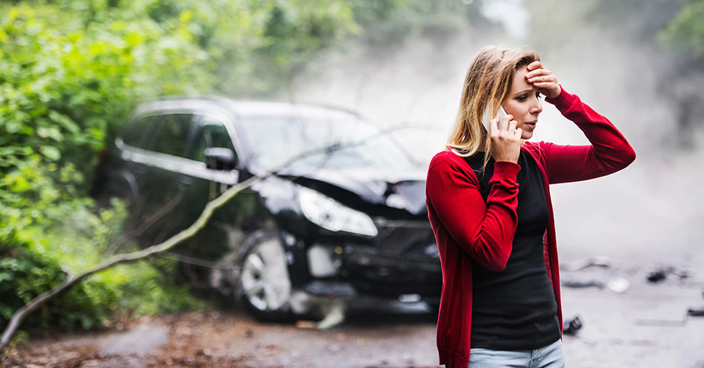 Woman on the phone looking distressed near a crashed vehicle on a wooded roadside with visible damage and smoke, illustrating insurance evaluation and Arizona Total Loss Threshold after a serious auto accident