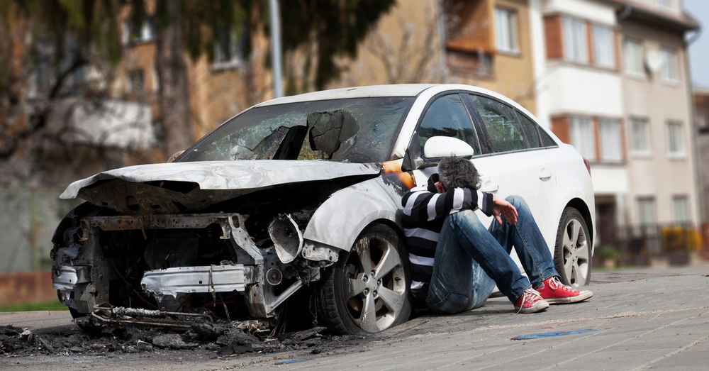 Burned and heavily damaged car after a severe crash with a distressed driver sitting beside it, illustrating insurance evaluation and Ohio Total Loss Threshold following a major auto accident