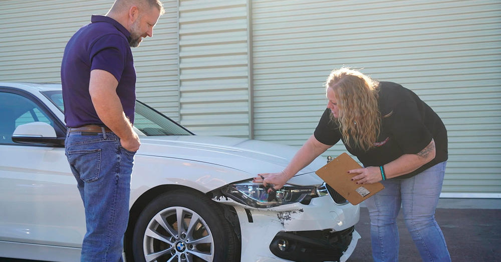 Insurance appraiser inspecting front-end headlight and bumper damage on a white sedan while discussing repairs with the owner, illustrating Total Loss Value Appraisal/Settlement after a vehicle accident